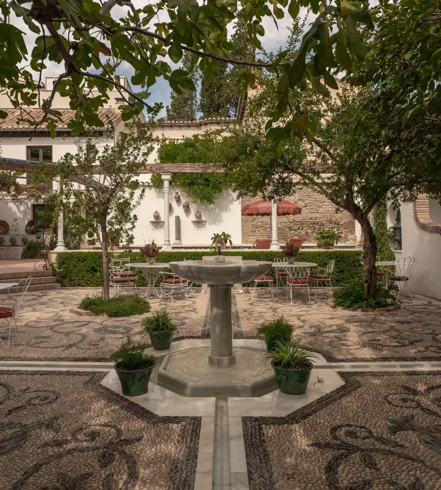 Patio andaluz con fuente de piedra, bancos y mesas entre &aacute;rboles y macetas