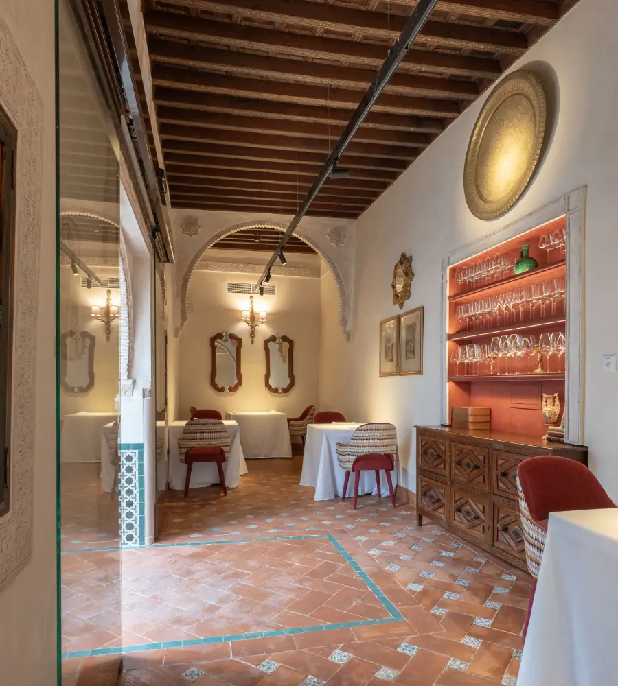 Comedor con mesas, arcos de piedra, azulejos y barra de cristal con luz c&aacute;lida.